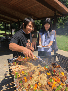 Ziyong Hong using tongs to grab a chicken skewer from the grill for Chia-Wei Hu who is using both hands to hold a paper plate