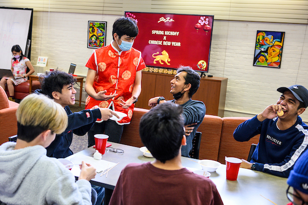 Standing and wearing a traditional Tang suit, Gan Zhao Yang, president of the Malaysian Student Association (MSA), talks with guests enjoying snacks as more than 120 people participate in a Lunar New Year celebration hosted by the Malaysian Student Association (MSA) in the Multicultural Center Lounge in the Red Gym (Armory and Gymnasium) at the University of Wisconsin-Madison on Feb. 5, 2022. A campus and county health mandate requires people to wear face mask indoors - except while eating or drinking - as the coronavirus (COVID-19) pandemic continues. (Photo by Jeff Miller / UW-Madison)