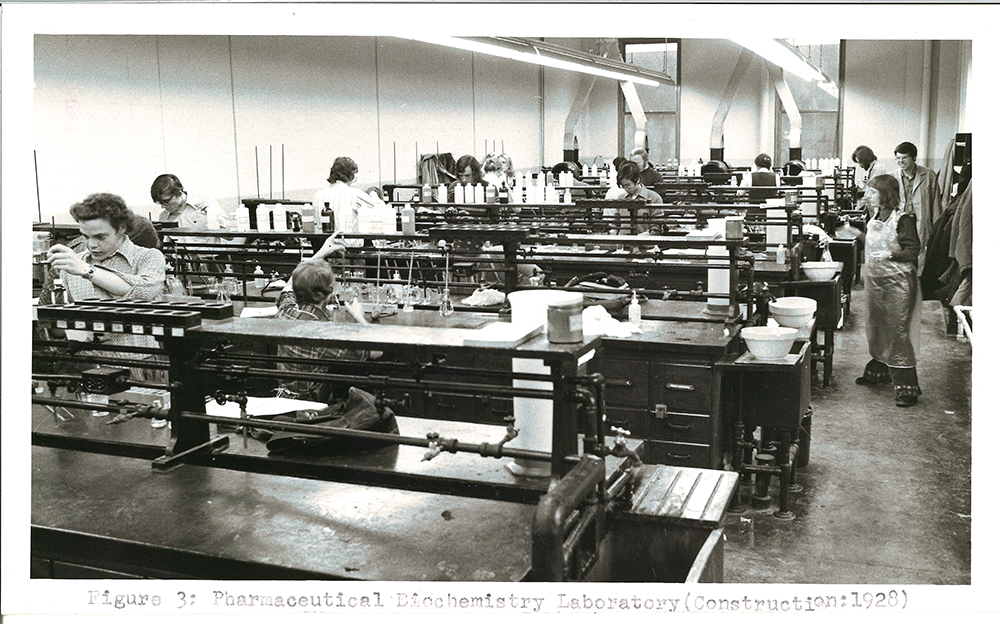 In a black-and-white photo, about a dozen students work at lab tables with various liquids. A woman on the right side of the photo wears a plastic apron.