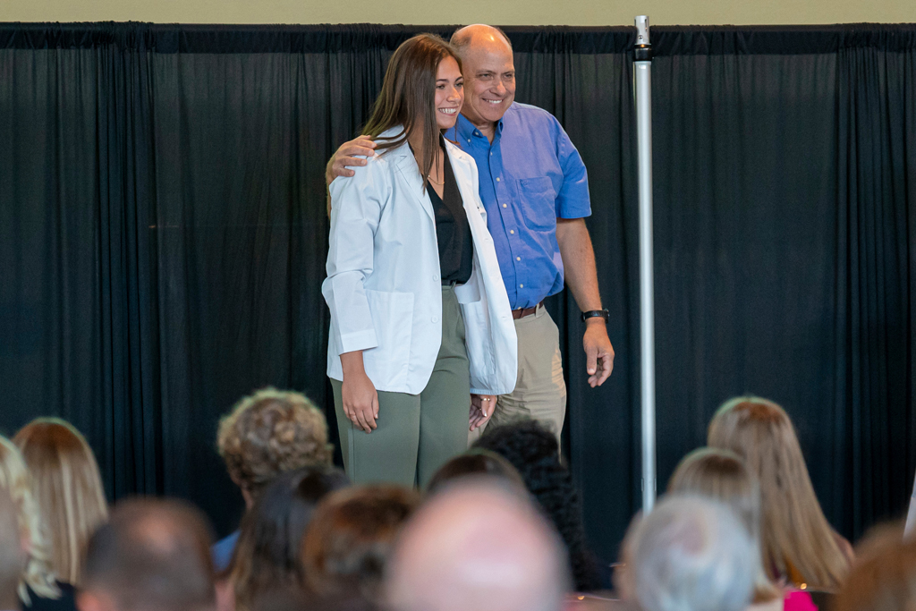 Student in white coat smiling with staff on-stage