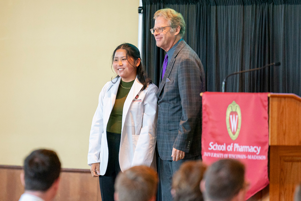 Student in white coat smiling with staff on-stage