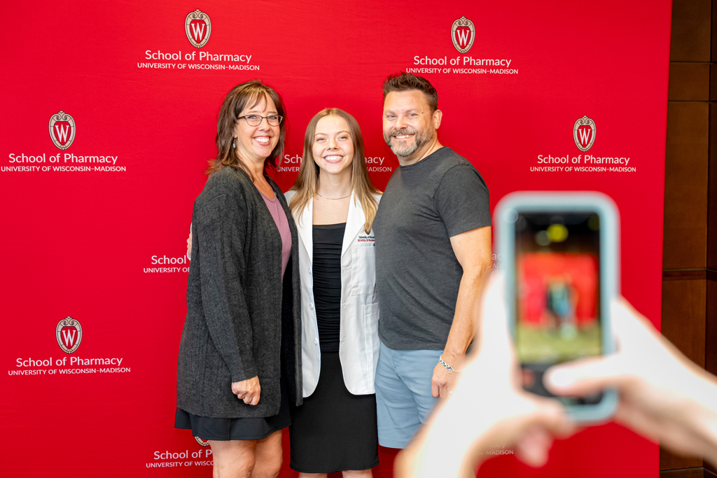 Student in white coat smiling with her parents in front of School of Pharmacy banner