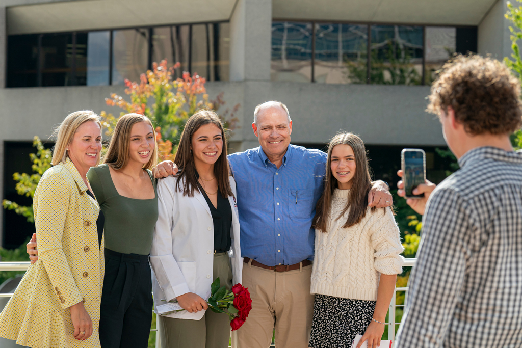 Student in white coat holding flowers and taking a photo with family