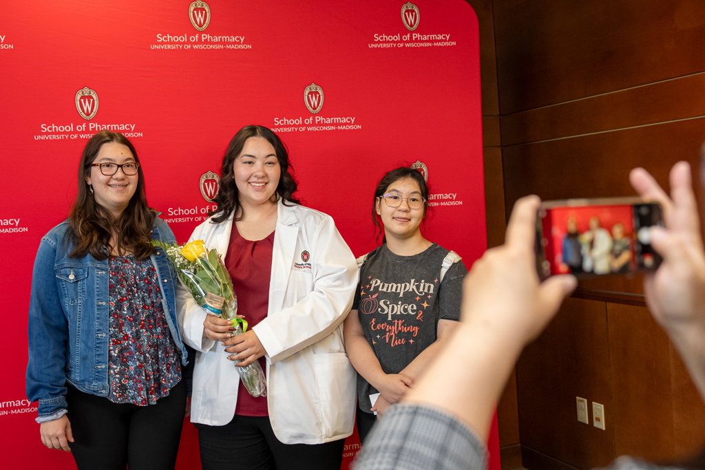 Student in white coat holding yellow rose with family in front of School of Pharmacy backdrop