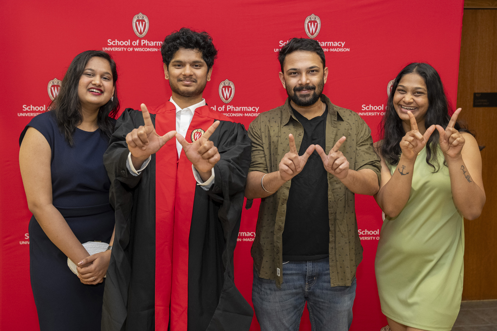 A graduate and family members pose in front of a red School of Pharmacy background