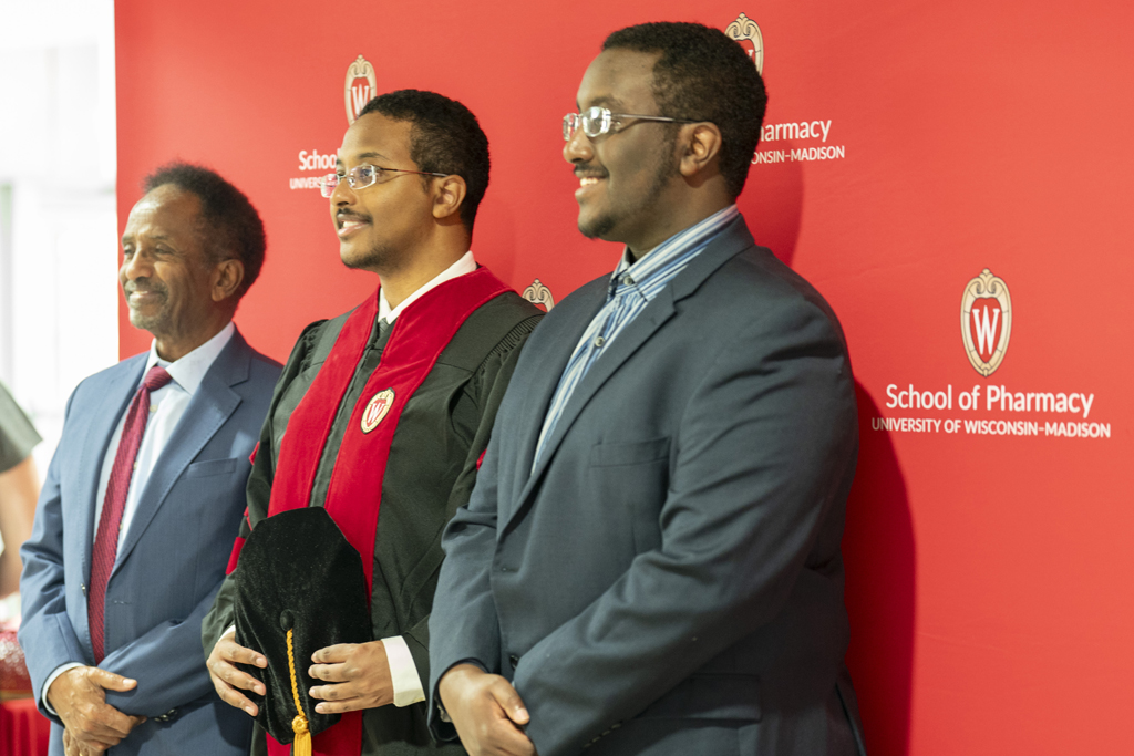 A graduate and family members pose in front of a red School of Pharmacy background