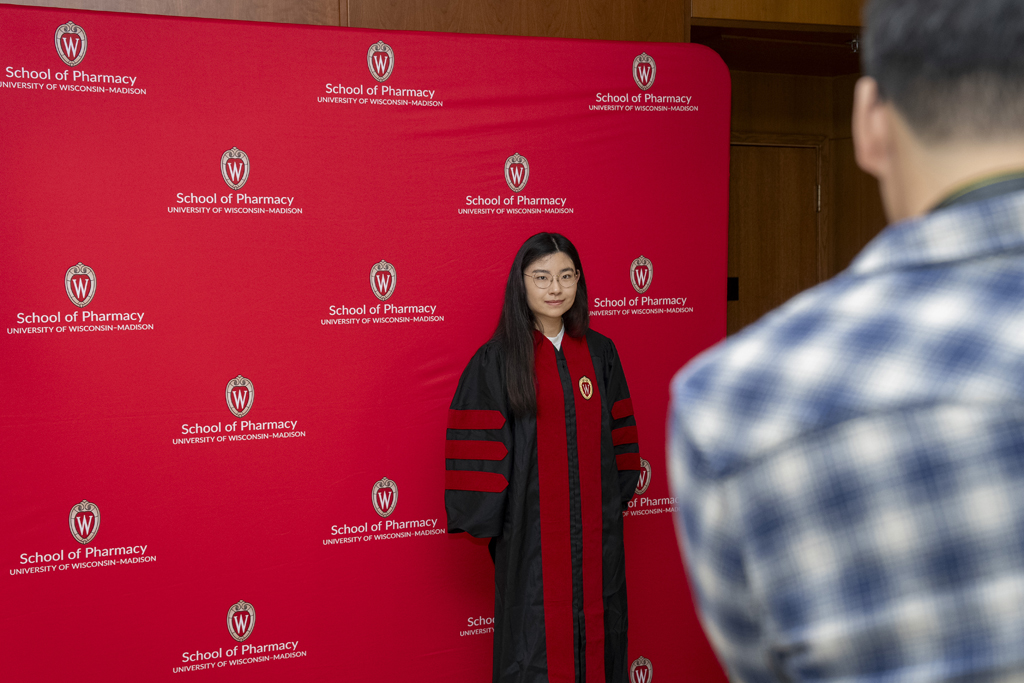 A graduate poses in front of a red School of Pharmacy background