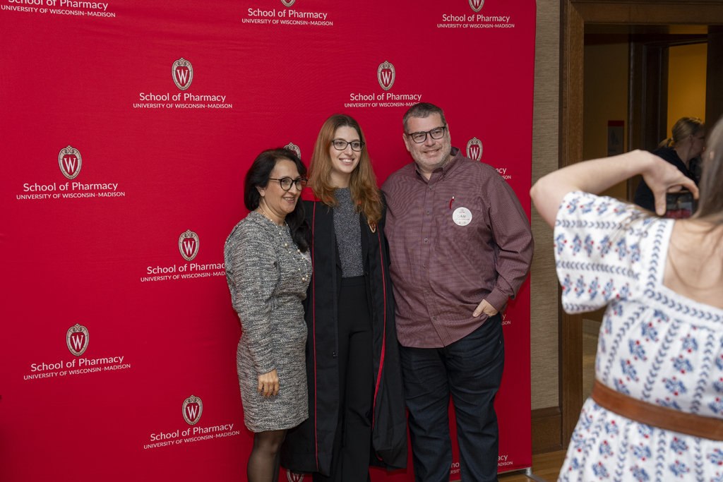 A graduate poses with his family in front of a red School of Pharmacy background