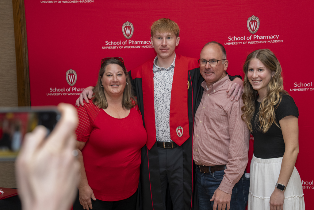 A graduate poses with his family in front of a red School of Pharmacy background