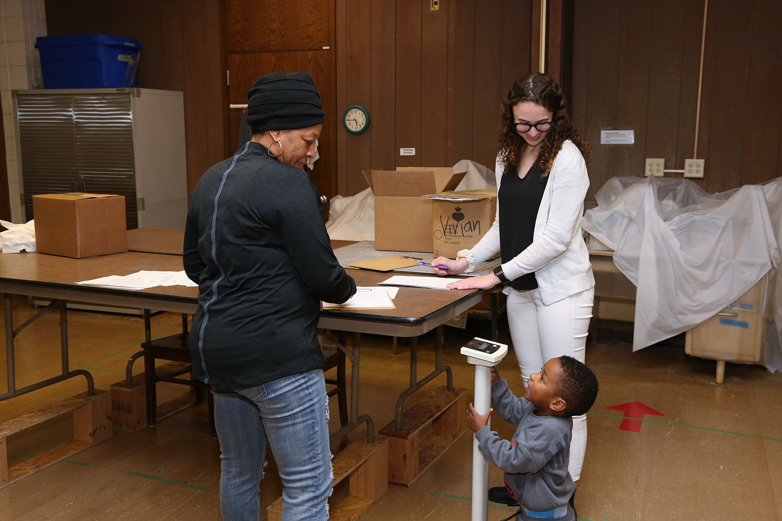 A participant speaks with a PharmD student at a table, with a young child standing on a scale nearby.