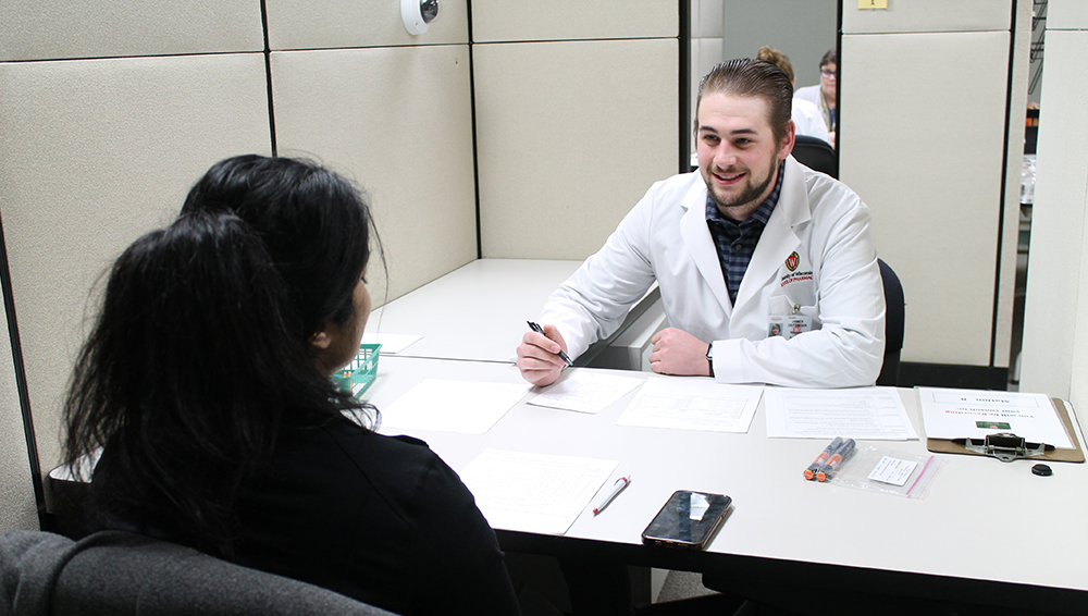 A student in a white coat speaks with a dark-haired person across a table.