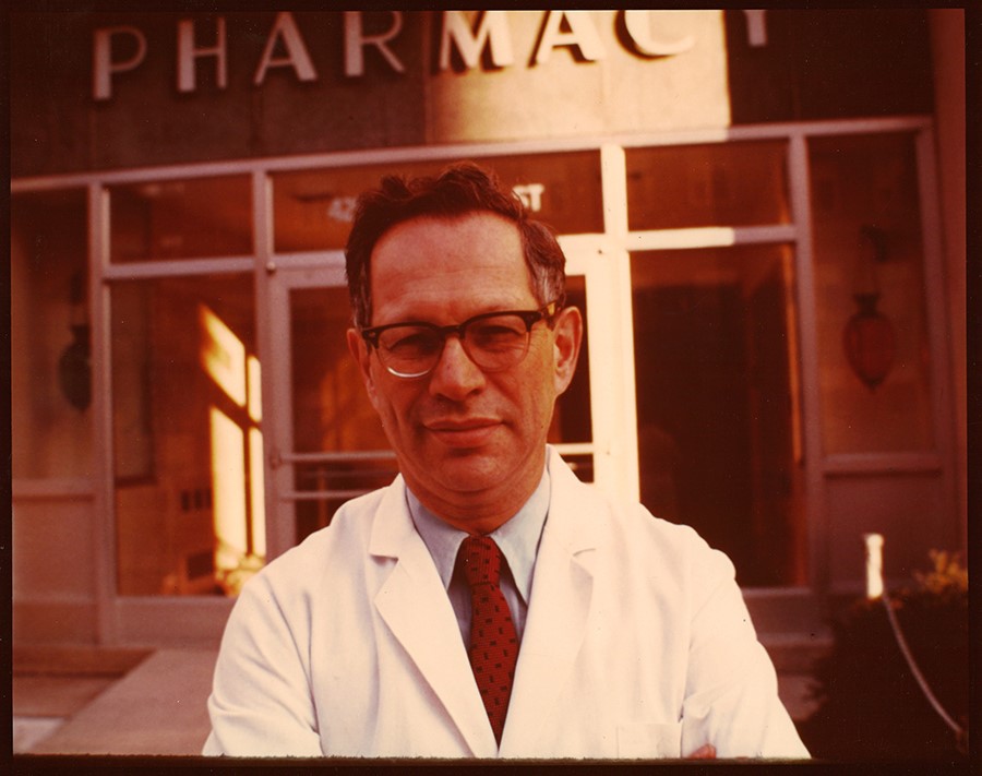 In an antique photo, David Perlman stands in front of the entrance to the School of Pharmacy wearing his white coat.