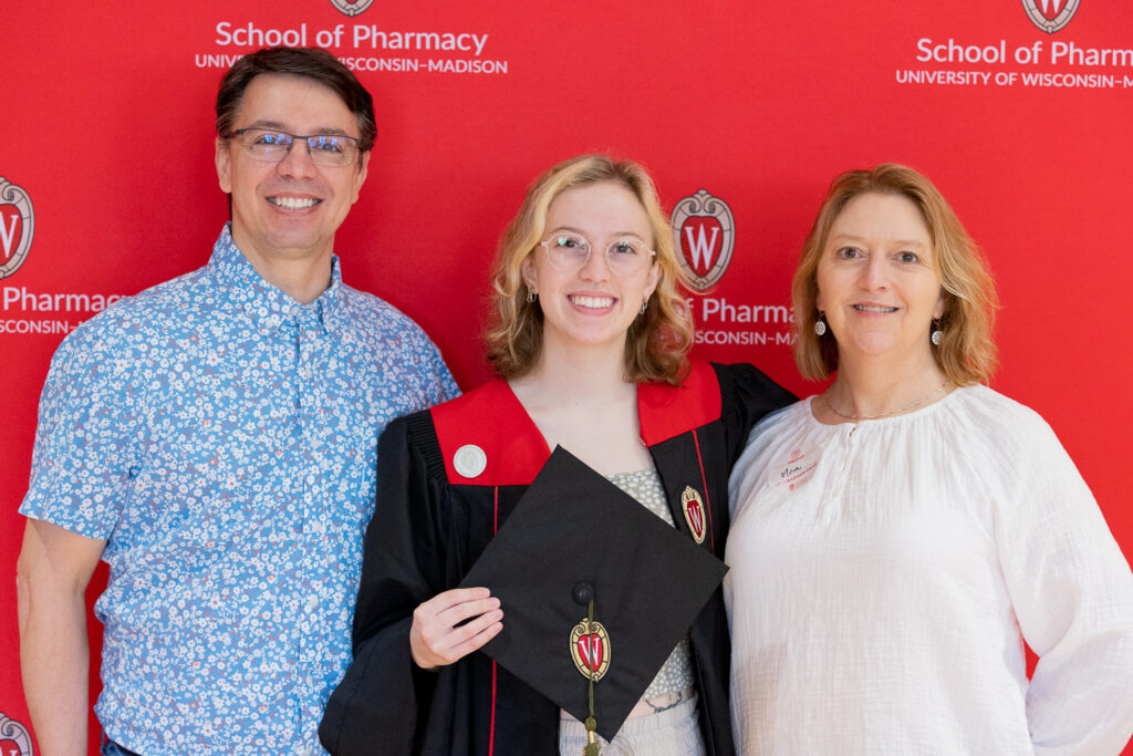A PharmTox graduate and her family pose in front of the red School of Pharmacy backdrop.