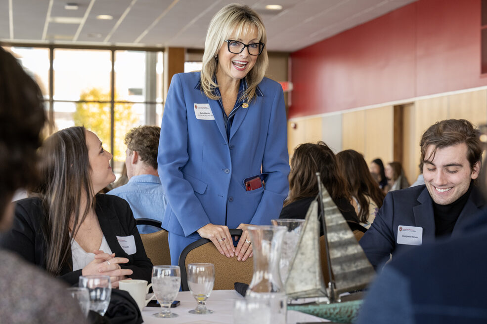 Beth Martin standing and resting hands against empty chair while laughing with seated students