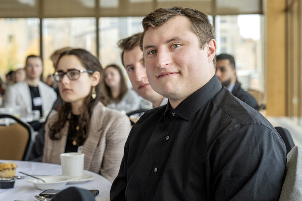 Camera focused on Kain Carstens listening to speaker with his brother and other students visible behind him out of focus