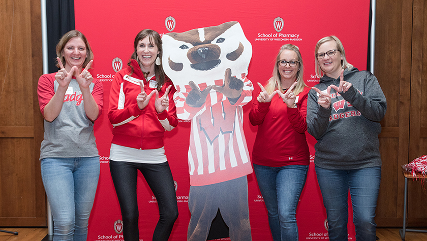 Four alumni stand with a cutout of Bucky in front of a red School of Pharmacy backdrop.