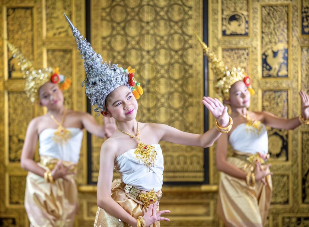 Three Thai women wearing ornate metallic tall hats and jeweled gold necklaces dance in front of an ornate screen
