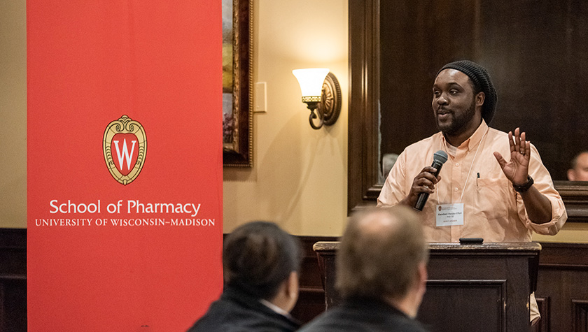 Kwadwo Owusu-Ofori speaking into a microphone in front of a UW-Madison School of Pharmacy banner