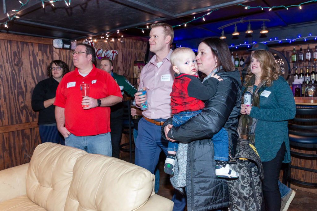 UW Pharm Alumni holding drinks and staring off-screen at at a TV displaying the Wisconsin vs Northwestern basketball game