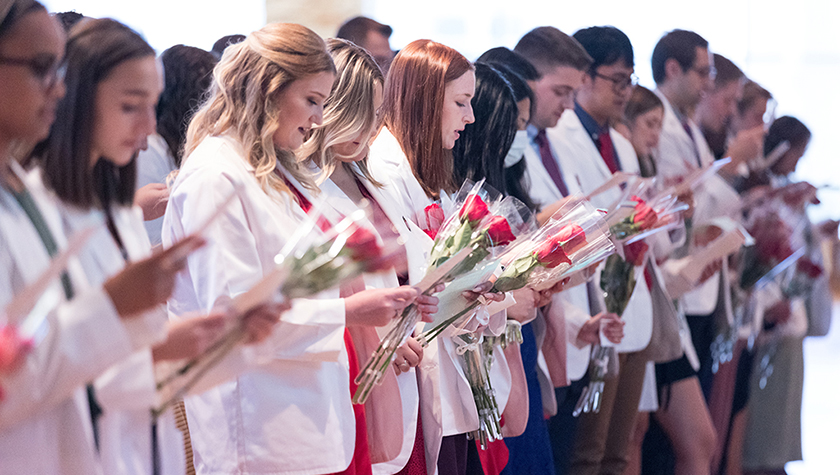 A row of PharmD students in white coats and holding red roses read from papers in their hands.