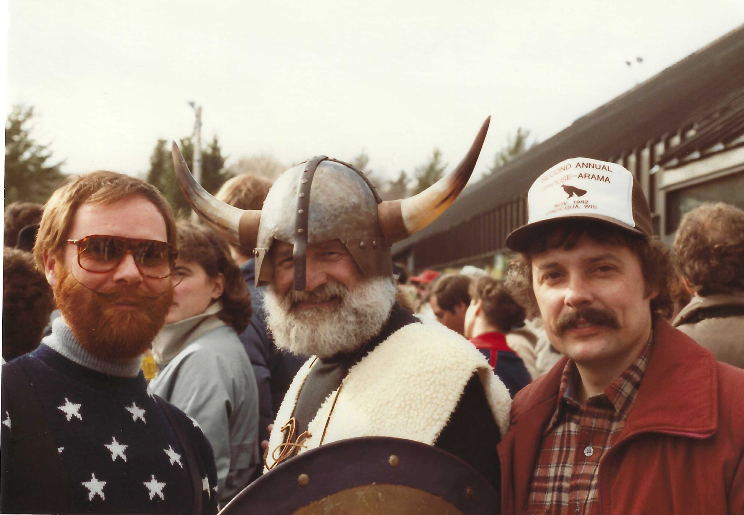 Three men are pictured outdoors in a crowd. The center man wears a Viking-style horned helmet and fur-lined garment. The left man has a red beard, sunglasses, and a star-patterned sweater. The right man wears a mustache, trucker hat, plaid shirt, and red jacket.