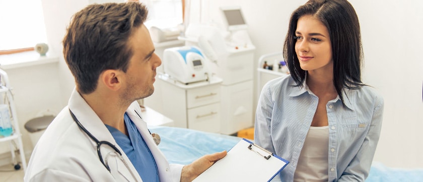A pharmacist with a clipboard sits with a patient to ask about side effects from her new medication