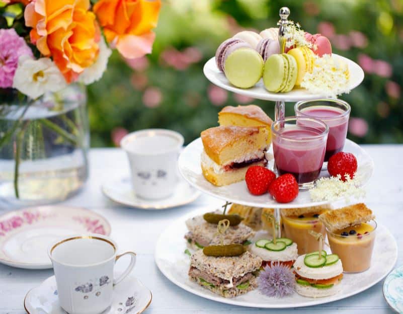 A colorful bouquet of flowers, tea cups, and a trivet with finger sandwiches, elegant pastries, puddings, and fresh strawberries sit on a white table