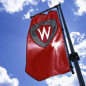 A "W" crest banner flies on Bascom Hill against blue sky and puffy clouds during spring.