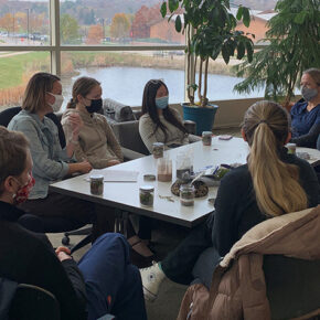 A group of students sitting around a table talking in a room full of windows.