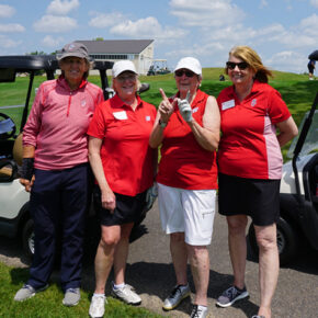 Four female alumni wearing red stand and smile in front of golf carts