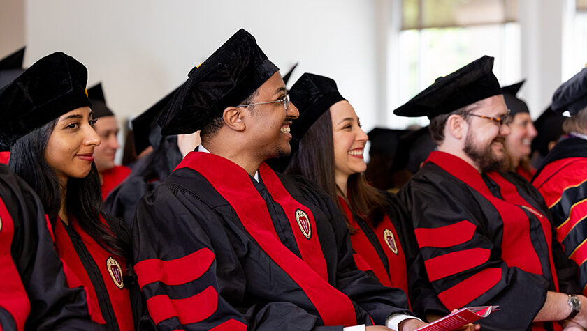 School of Pharmacy graduates in hats and robes smile while listening to a speaker.