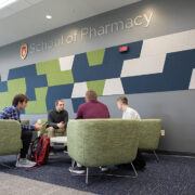 Comfortable lounge-style seating in front of a blue-and-green tiled wall with the "School of Pharmacy" logo on it. Students study in the seats.