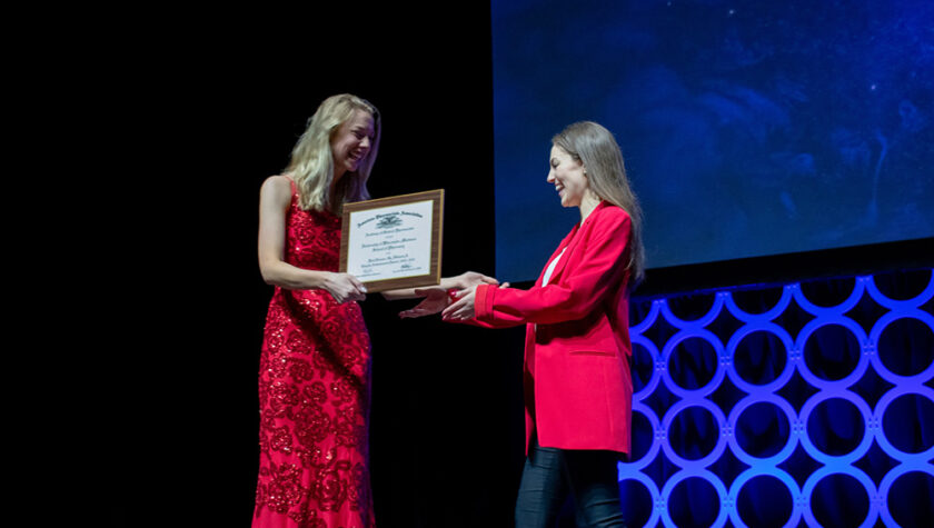 Lauren Glaza accepts an award onstage from a woman in a red dress.