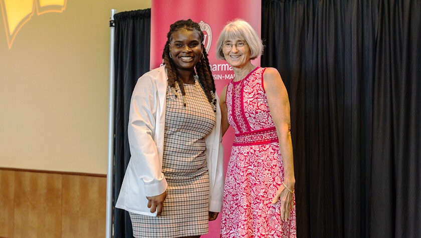 A PharmD student poses in her white coat next to Professor Mara Kieser, in a red dress.