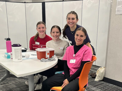 PharmD students Amalia Fox, Hannah Whitney, Chaise Pucek, and Nicole Ulbricht pose behind a white folding table with vaccination supplies on it.