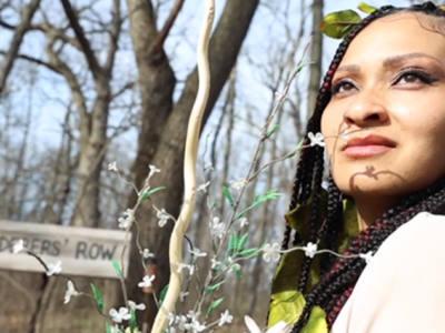 Thi Le, wearing leaves in her hair, looks back over her shoulder next to a sign reading "murderers' row."