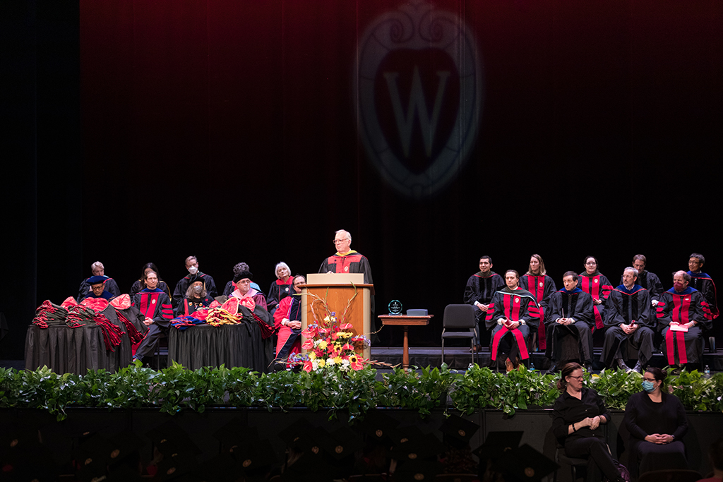 Pan out shot of professors sitting onstage with a speaker at the podium and hoods laying on tables to the left