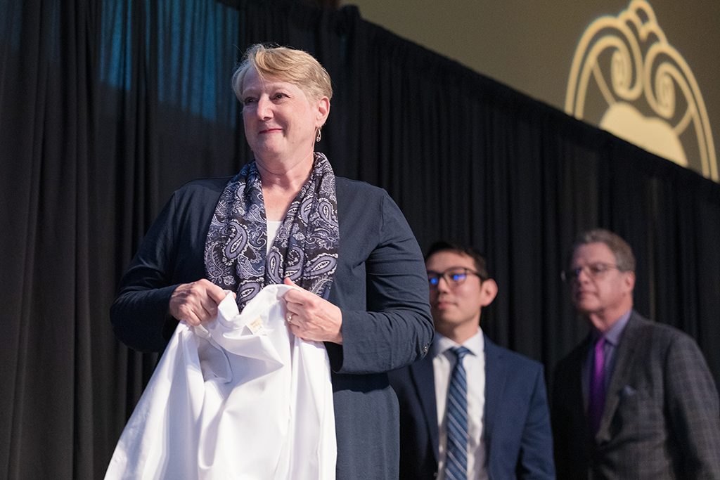 Associate Professor Karen Kopacek holds a white coat for a student.