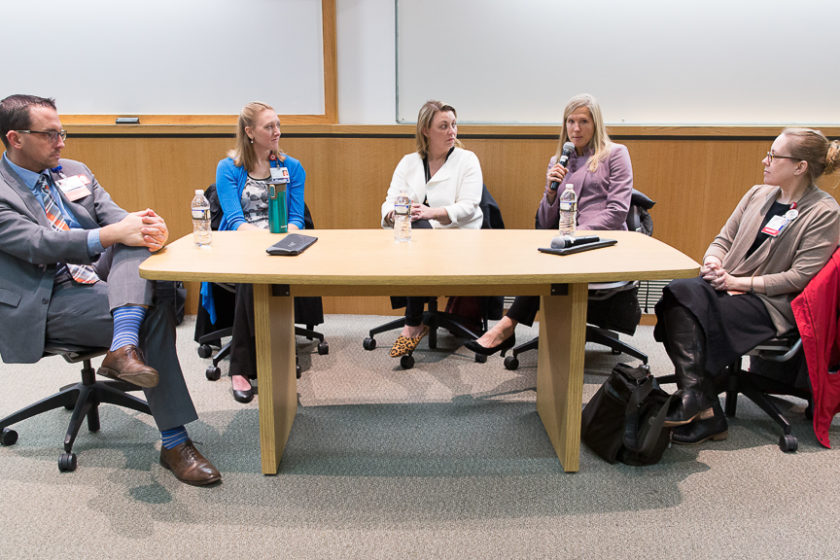 Panel of people sitting behind table with one of them speaking into microphone